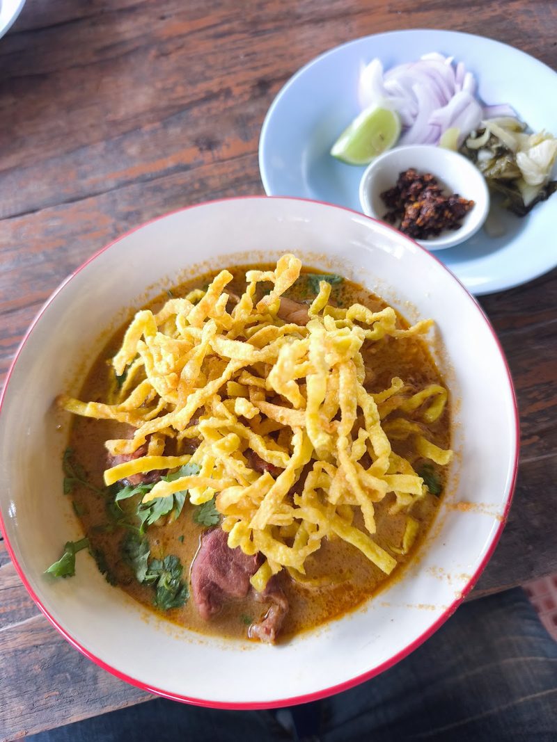 ข้าวซอยโรยเส้นกรอบในชามขาว มีจานหอมแดง ผักกาดดอง และมะนาววางข้าง ๆ
