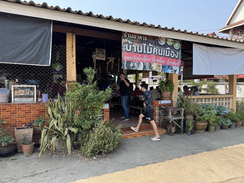 หน้าร้านบ้านไม้คนเมือง ป้ายชื่อร้านเขียนว่า ขนมจีนน้ำเงี้ยว ก๋วยเตี๋ยว ข้าวซอย มีกระถางต้นไม้เรียงริมทาง